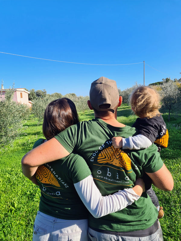 Woman, Man and Toddler as seen from behind with Obìa Olive Oil T-shirts in an olive grove